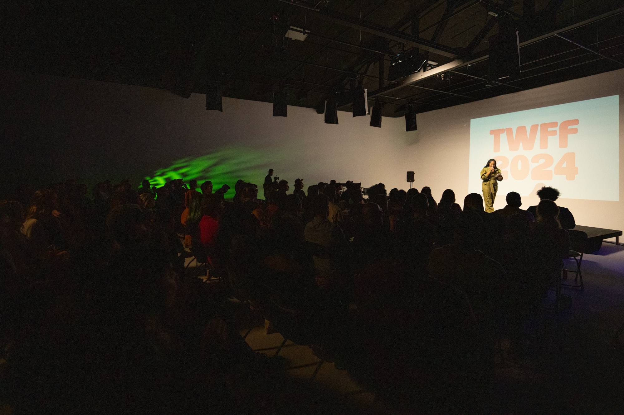 A dark room full of audience members focus on one person standing on stage with a microphone.