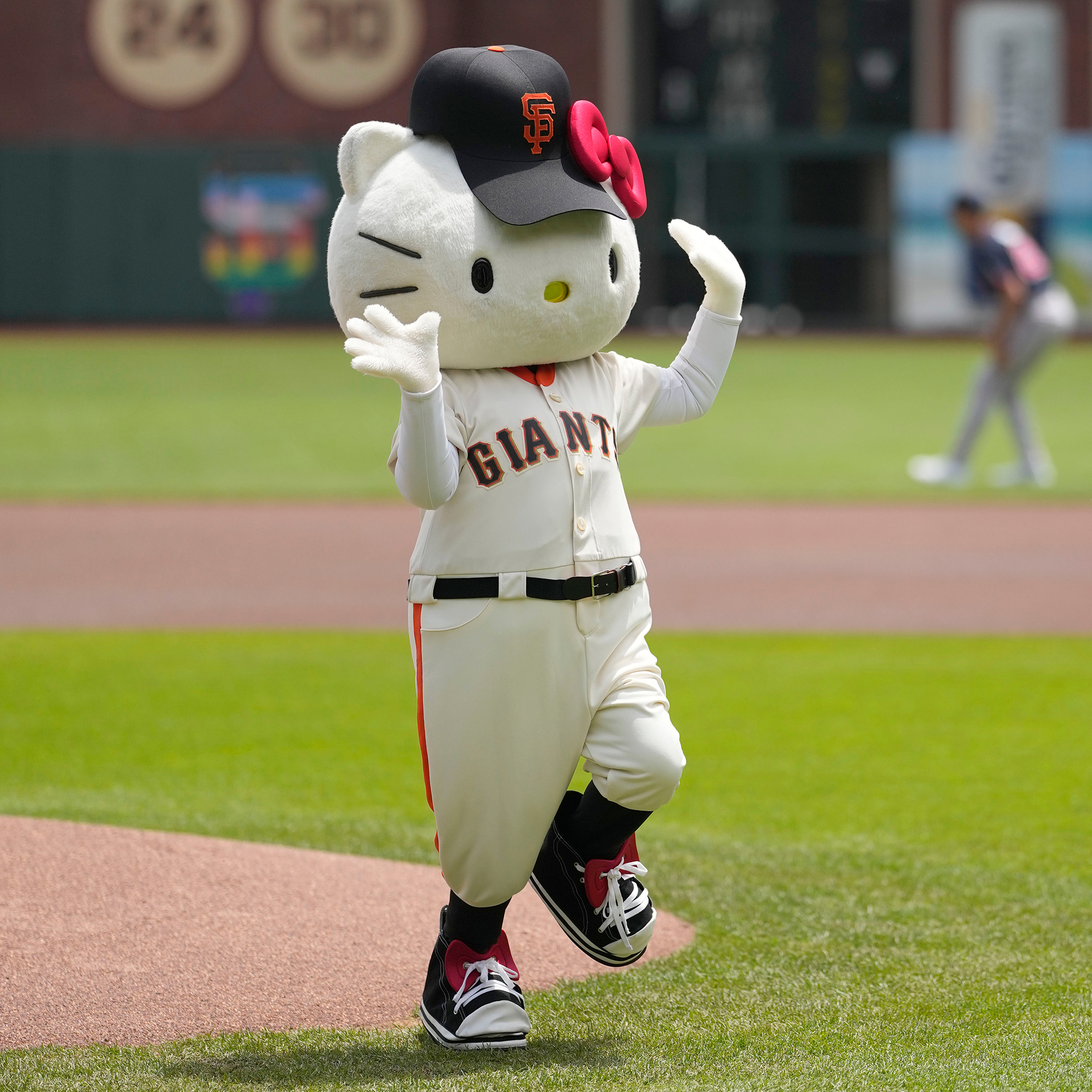 A person in a large white kitten outfit wearing a cap and jersey for the Giants baseball team waves with both hands on the baseball field