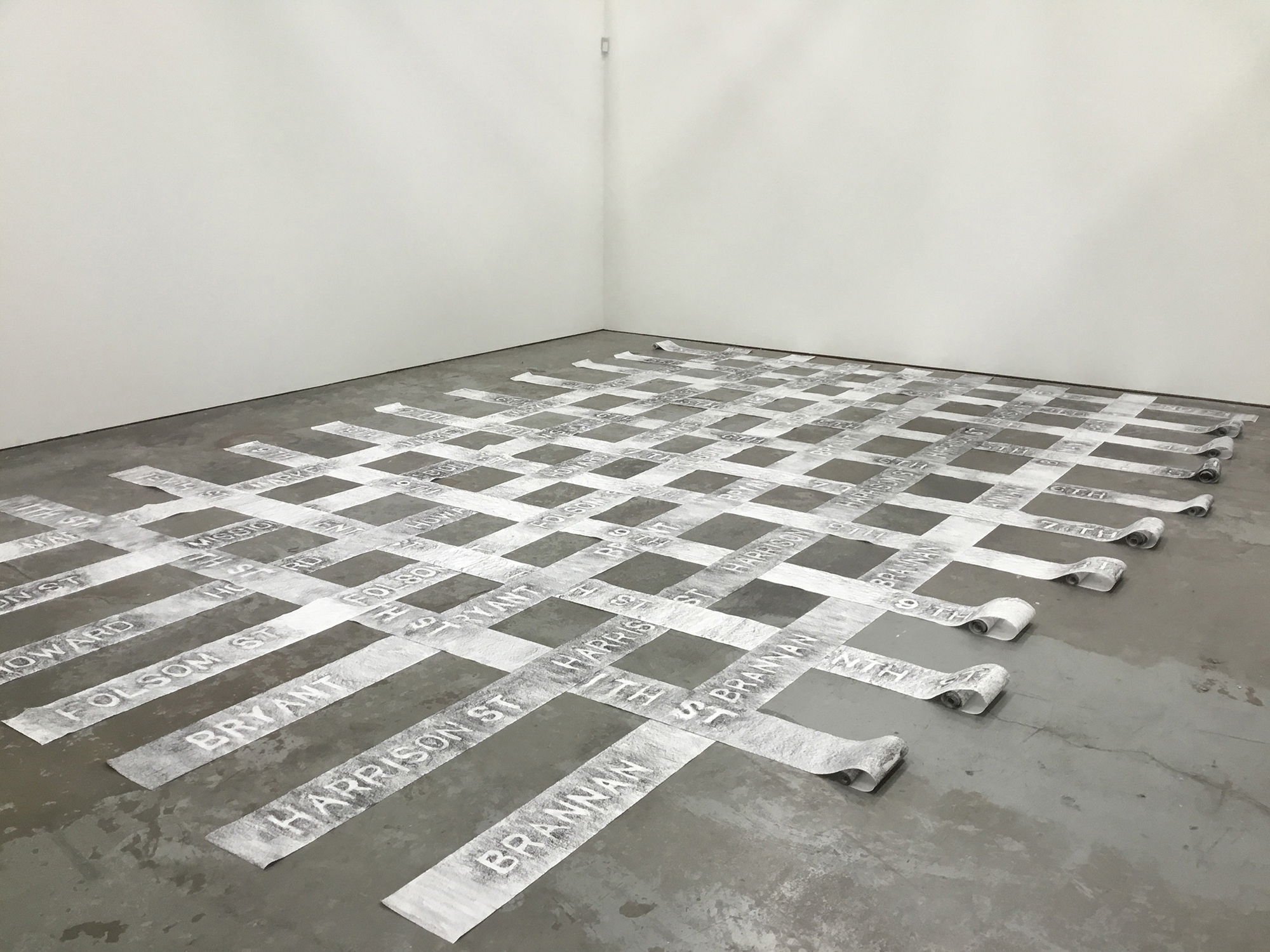 A grid of paper strips, covered in impressions of city street names from the sidewalk, lays on a concrete floor