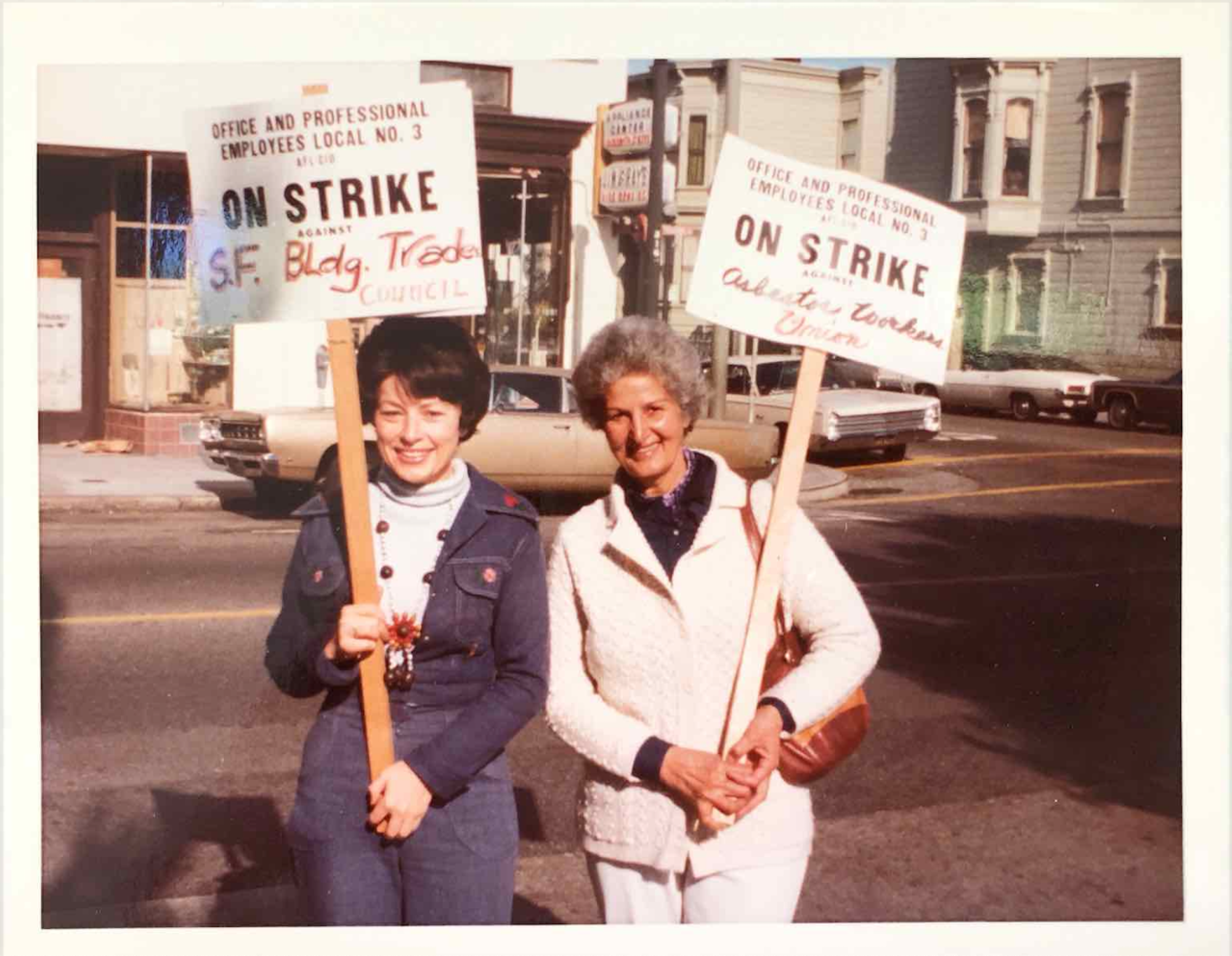 Two women in 1970s-era clothing stand side by side, smiling and holding placards during a protest.