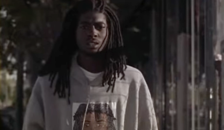 A young Black man with shoulder length dreadlocks stands in the street, looking concerned. He is wearing a grey hoodie.