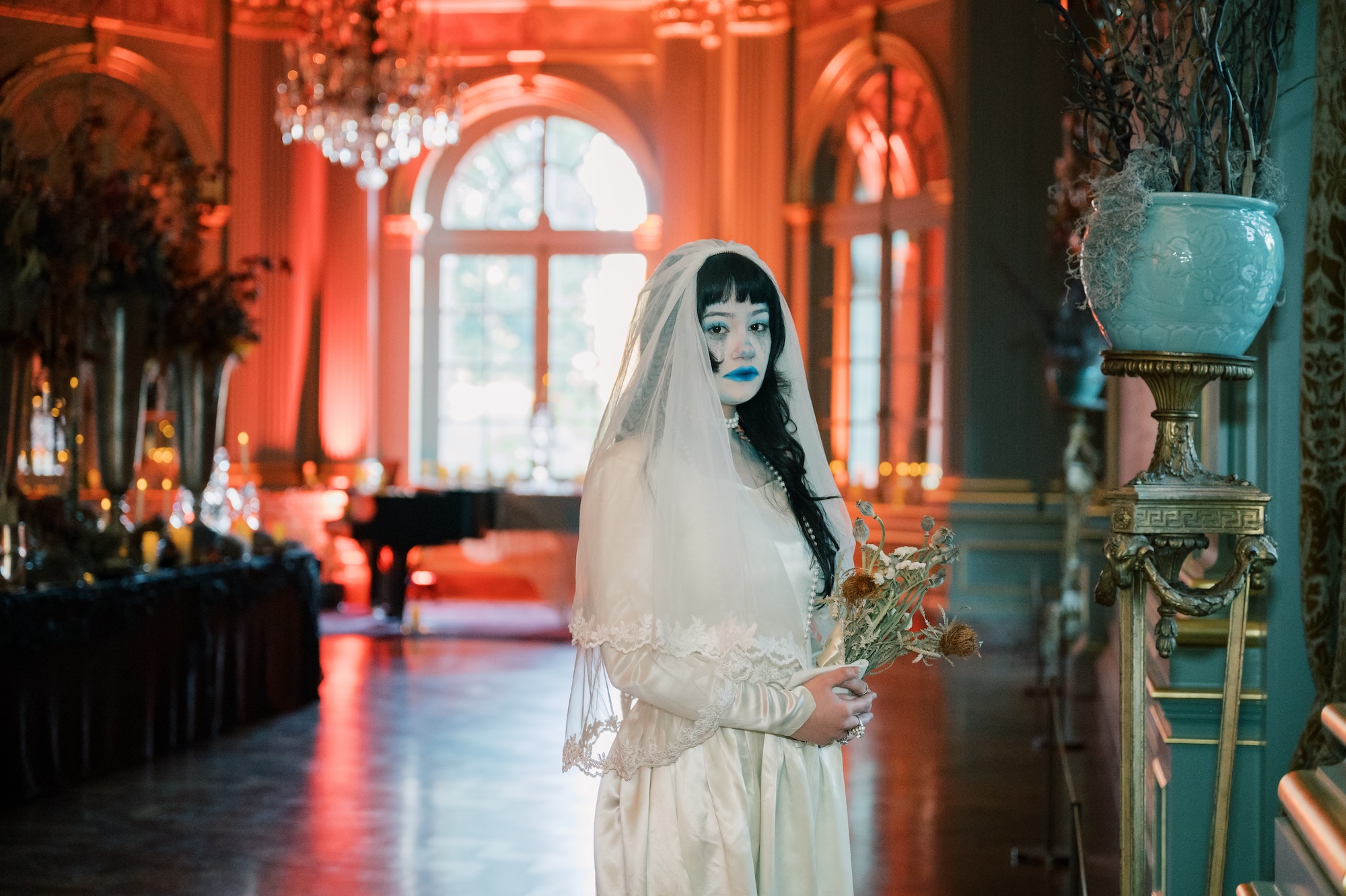 A woman wearing a white dress, veil and ghostly make-up stands in a grand hall, lit in red. She is holding dried flowers.