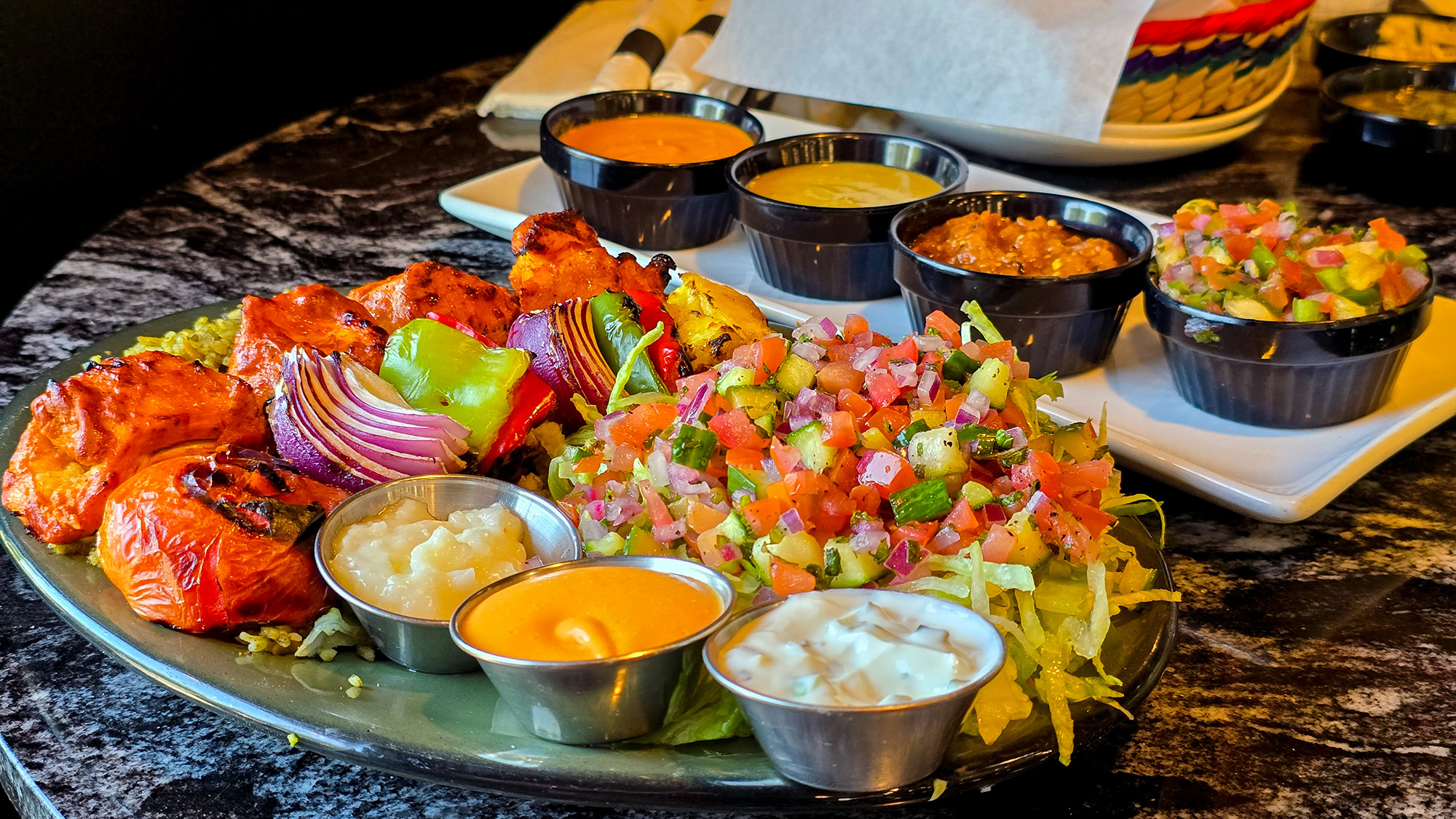 Chicken kebab, grilled tomatoes, cucumber salad and various sauces on a plate.