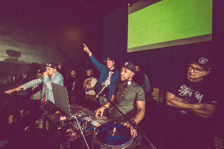 A group of people stand on stage, DJing, rapping or dancing