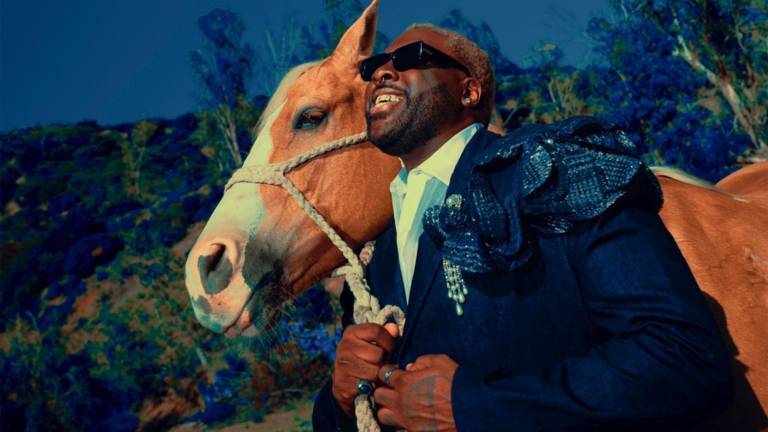 A young Black man wearing a deep blue suit holds a tan horse by the reins and looks up toward the sky, wearing sunglasses