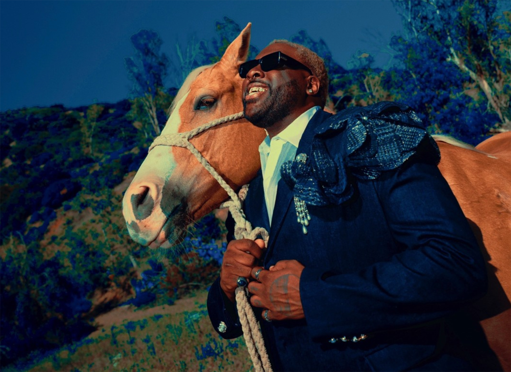 A young Black man wearing a deep blue suit holds a tan horse by the reins and looks up toward the sky, wearing sunglasses