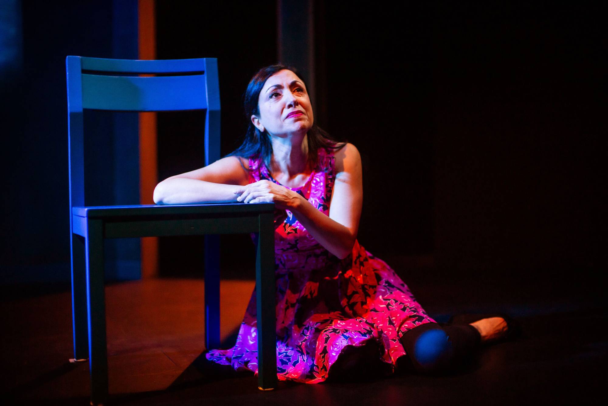 A woman in a pink dress sits on the floor, leaning against a chair, with tears rolling down her face.