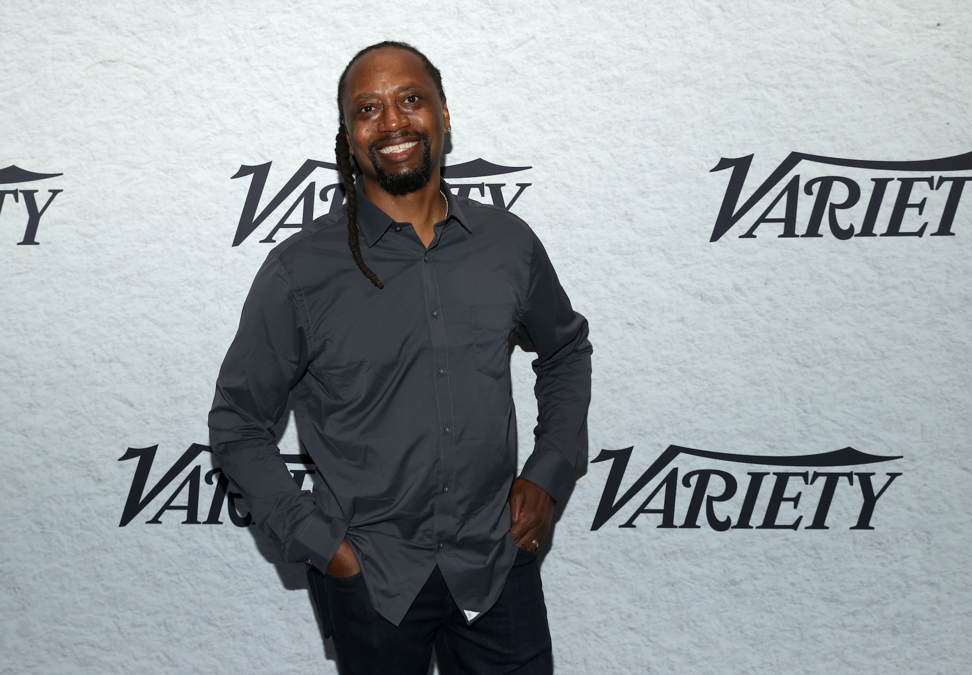 A smiling Black man wearing casual grey shirt and jeans stands in front of a white wall featuring the 'Variety' magazine logo.