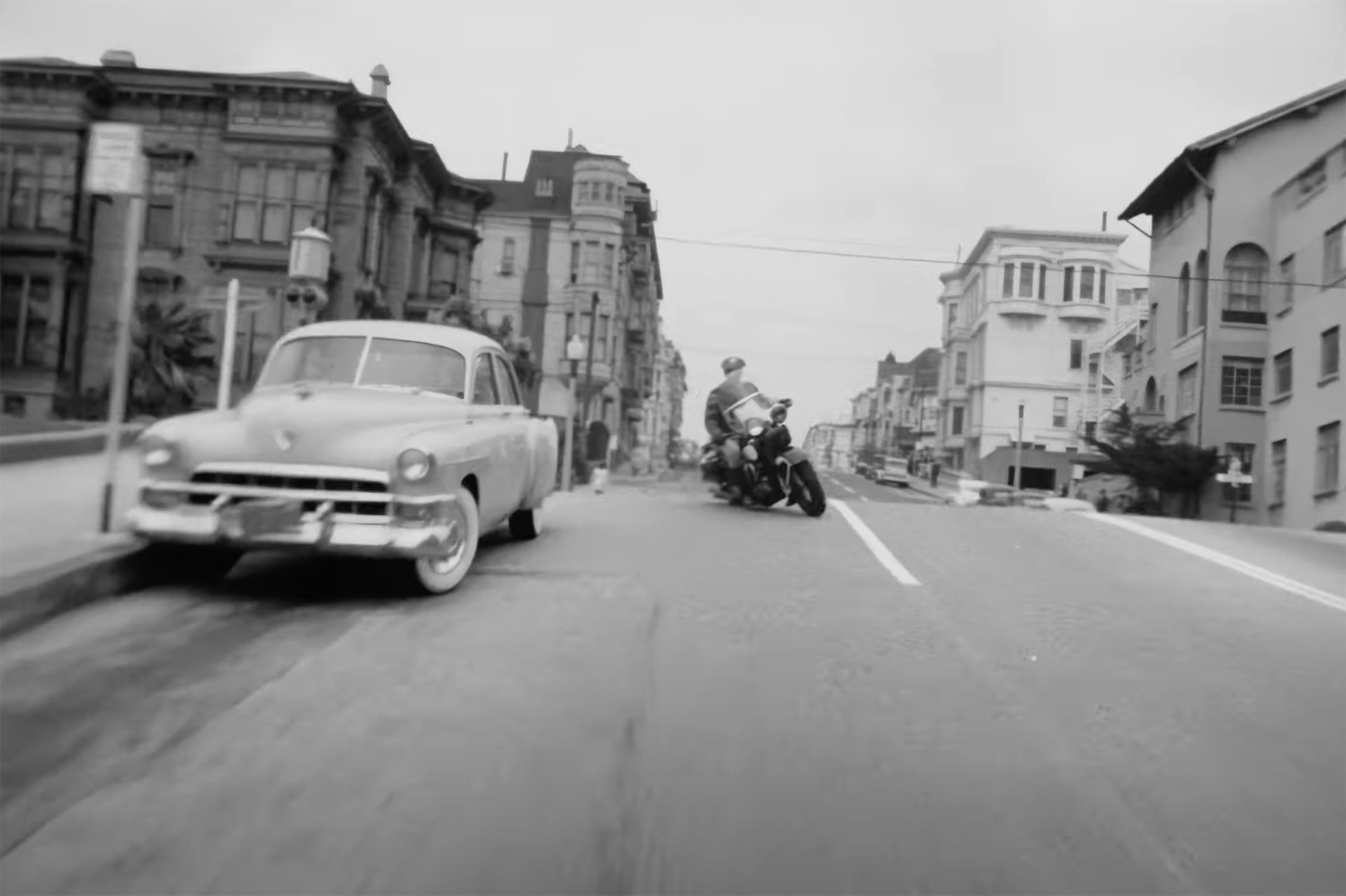 A police officer on a motorcycle leans into a corner turn with a large Victorian building in the background, in a still from a black-and-white movie
