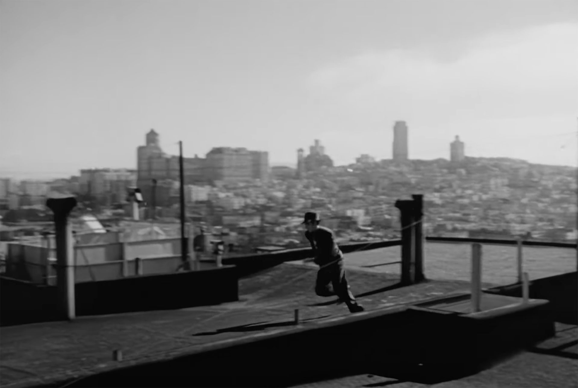 A man in a suit and fedora runs across a rooftop with a city skyline in the background, in a still from a black-and-white movie