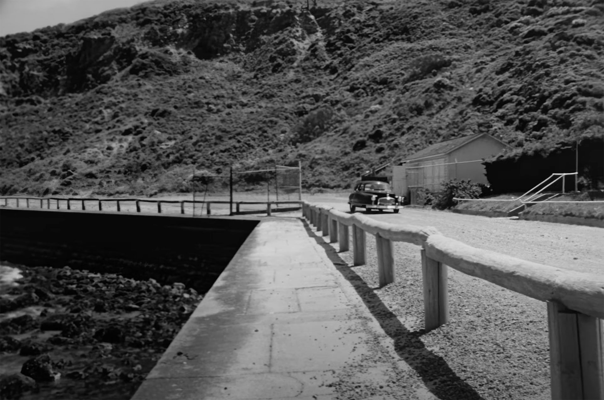 A large sedan drives along a modest road, lined with a waist-high fence next to a body of water, in a still from a black-and-white movie