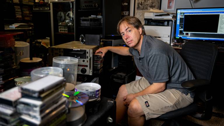 A man in his 50s wearing a polo shirt and beige shorts sits at a cluttered desk, his arm leaned upon a vintage analog tape machine