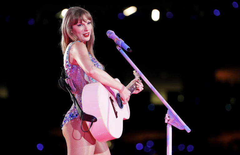 A white blond woman performs on a dark stage with a guitar. She is wearing a bejeweled body suit.