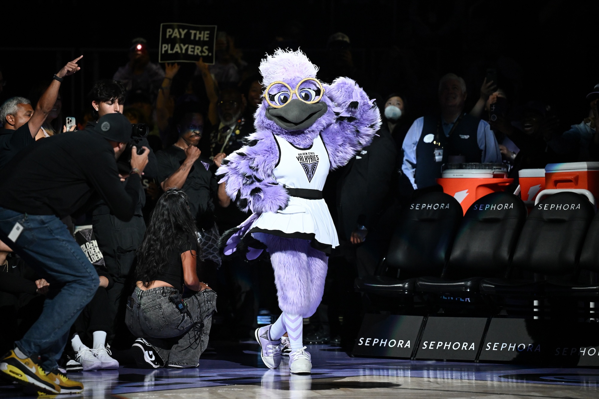 A sports mascot on the court strikes a pose. She is a purple bird decked out in a short white dress and gold eye glasses.