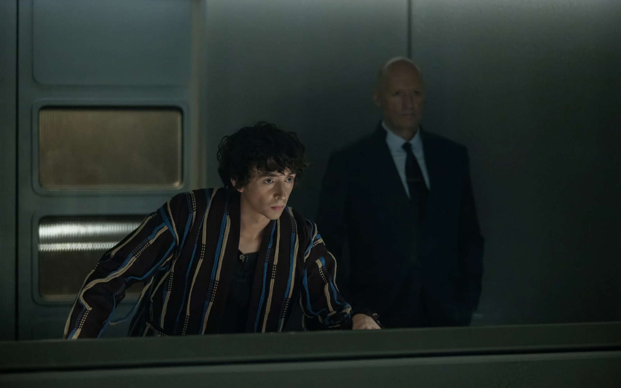 A young white man wearing a dressing gown leans forward over a console. Behind him stands an impassive bald white man wearing a formal suit.