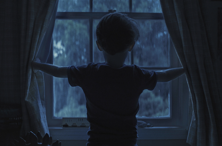 A small boy, viewed from behind, in dim morning light stands facing a window, stretching the curtains open.