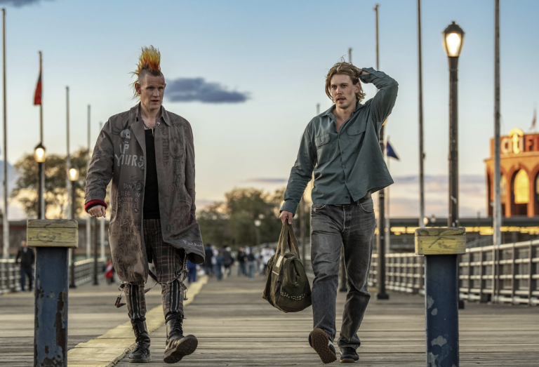 A white male punk rocker with a mohawk walks along a pier with a white man in his 30s. They are both in a scuffed up state.