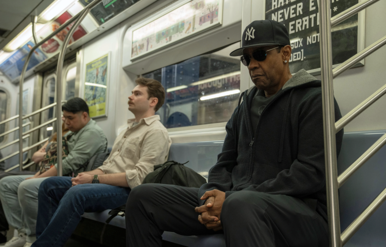 A Black man wearing sunglasses and a baseball cap sits on the subway, looking serious. There are two male passengers near by, paying him no mind.