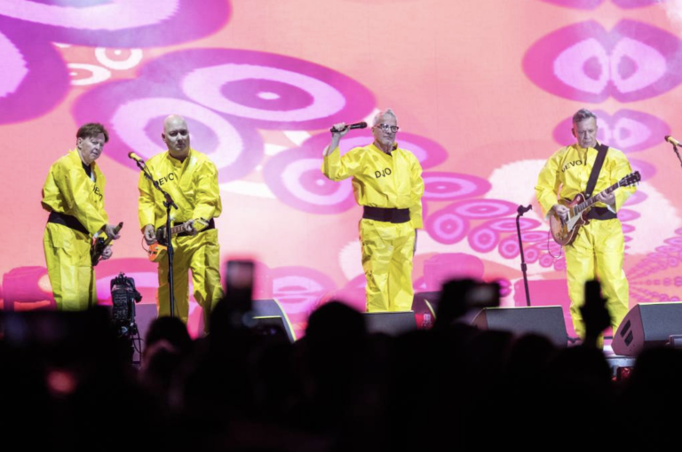 Four men wearing yellow jumpsuits perform music on stage in front of a pink backdrop.
