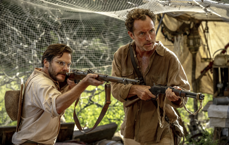 Two bedraggled and stressed-looking men wearing 1920s casual attire hold rifles aloft underneath a canopy of white netting. Jungle background.