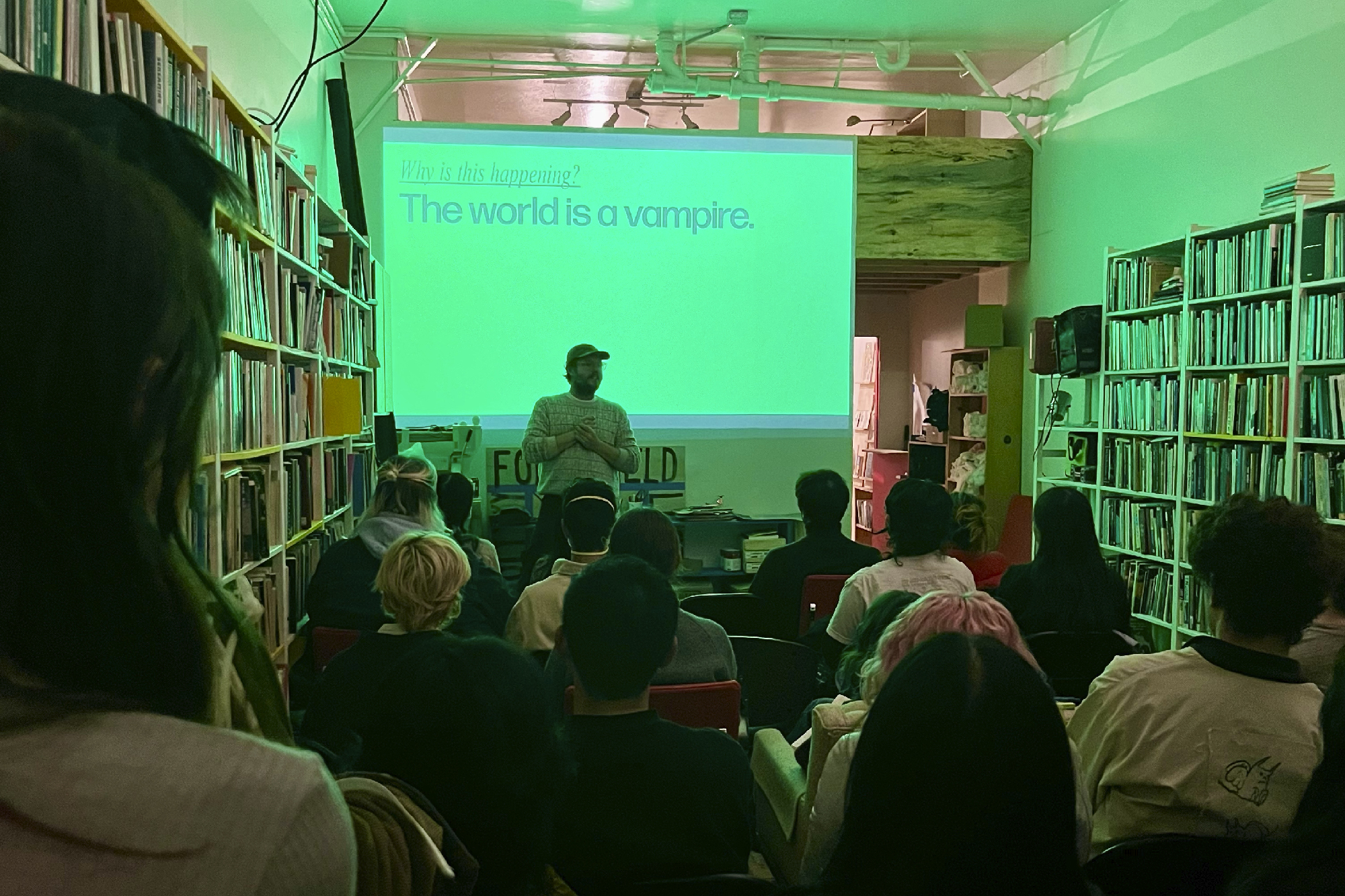 person in front of crowd in book-lined space with 'why is this happening? the world is a vampire' projected behind
