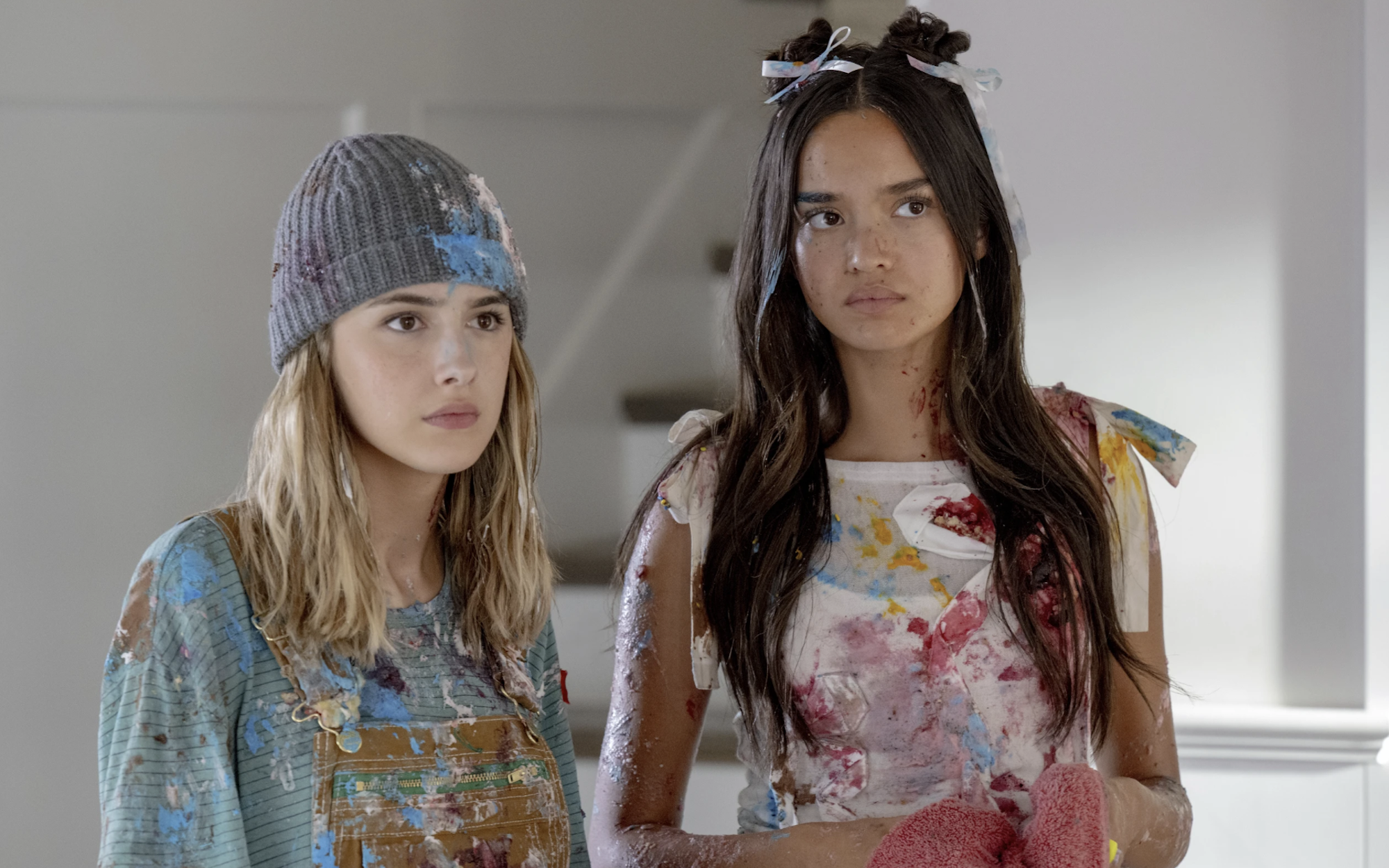 A white girl with scruffy blond hair and a taller, slender multiracial girl with long brown hair stand indoors together looking unimpressed. They are both covered with paint spatters. 
