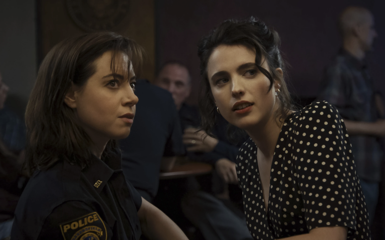 Two white women lean towards each other in a room busy with other people. The one on the left is wearing a police uniform. The one on the right is wearing a black dress with white polka dots on.