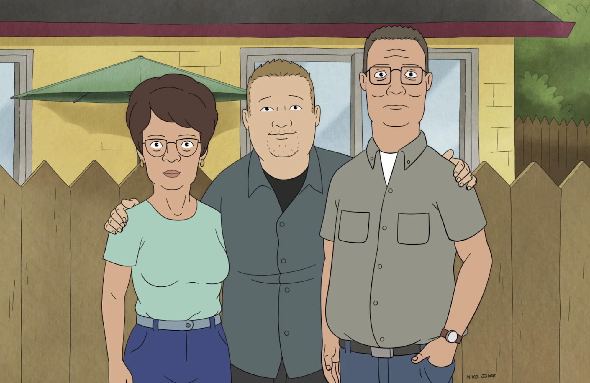 A still image from an animated TV show, showing a young man with his arms around an older woman and man. They are all standing in front of a brown fence around a yellow house.