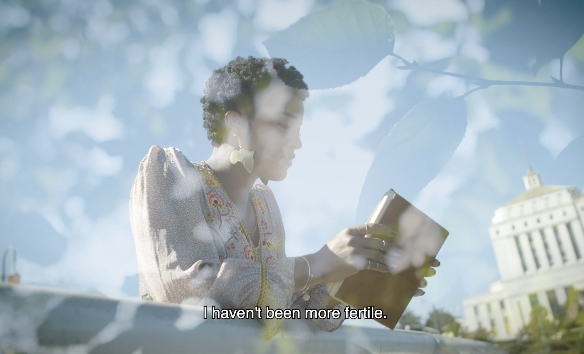 A Black woman with cropped hair leans on a railing, while holding a brown leather bound book. She is wearing a peasant dress.