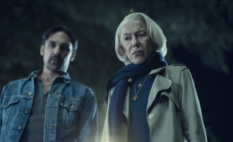 A senior woman with white hair wearing a beige raincoat stands looking down at the ground. Behind her stands a younger white man wearing casual shirt and denim jacket. He is also looking down at something.