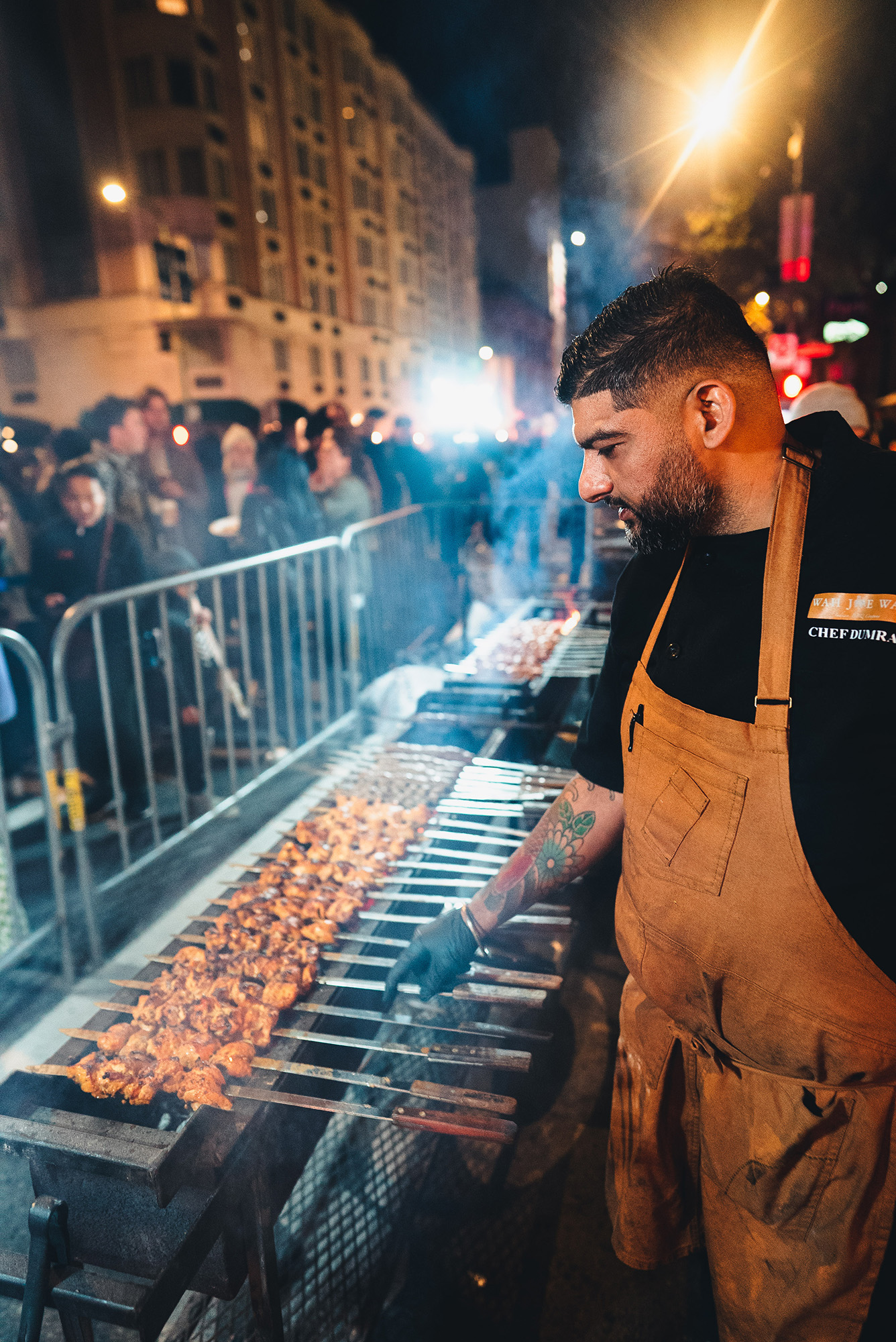 A chef grilling meat skewers outdoors while a crowd looks on.