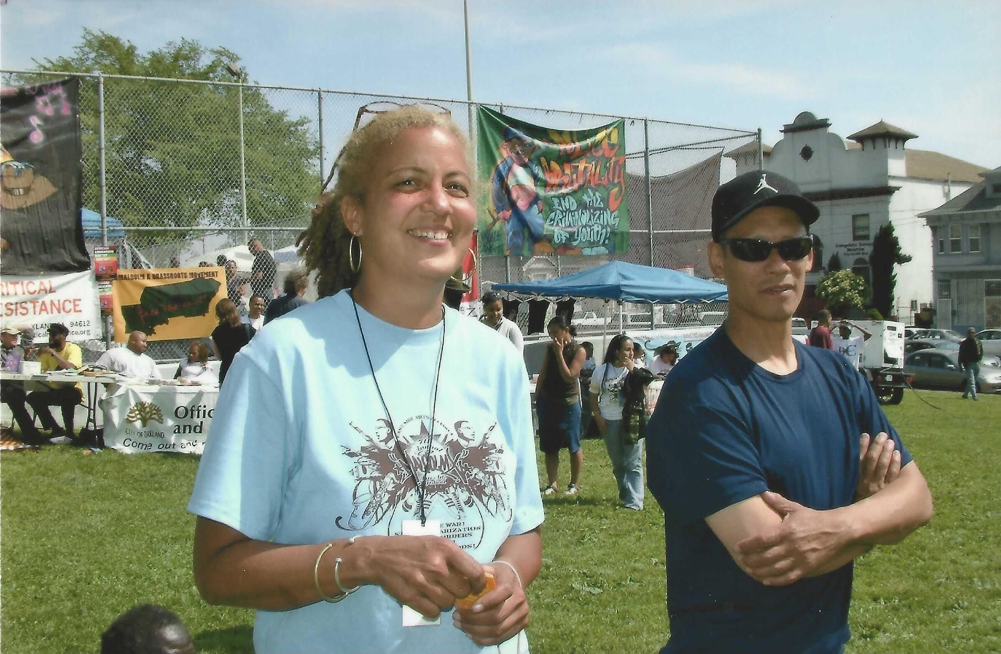 A photo of a woman in a light blue shirt and a man in a black shirt, sun glasses and hat standing in a park. 