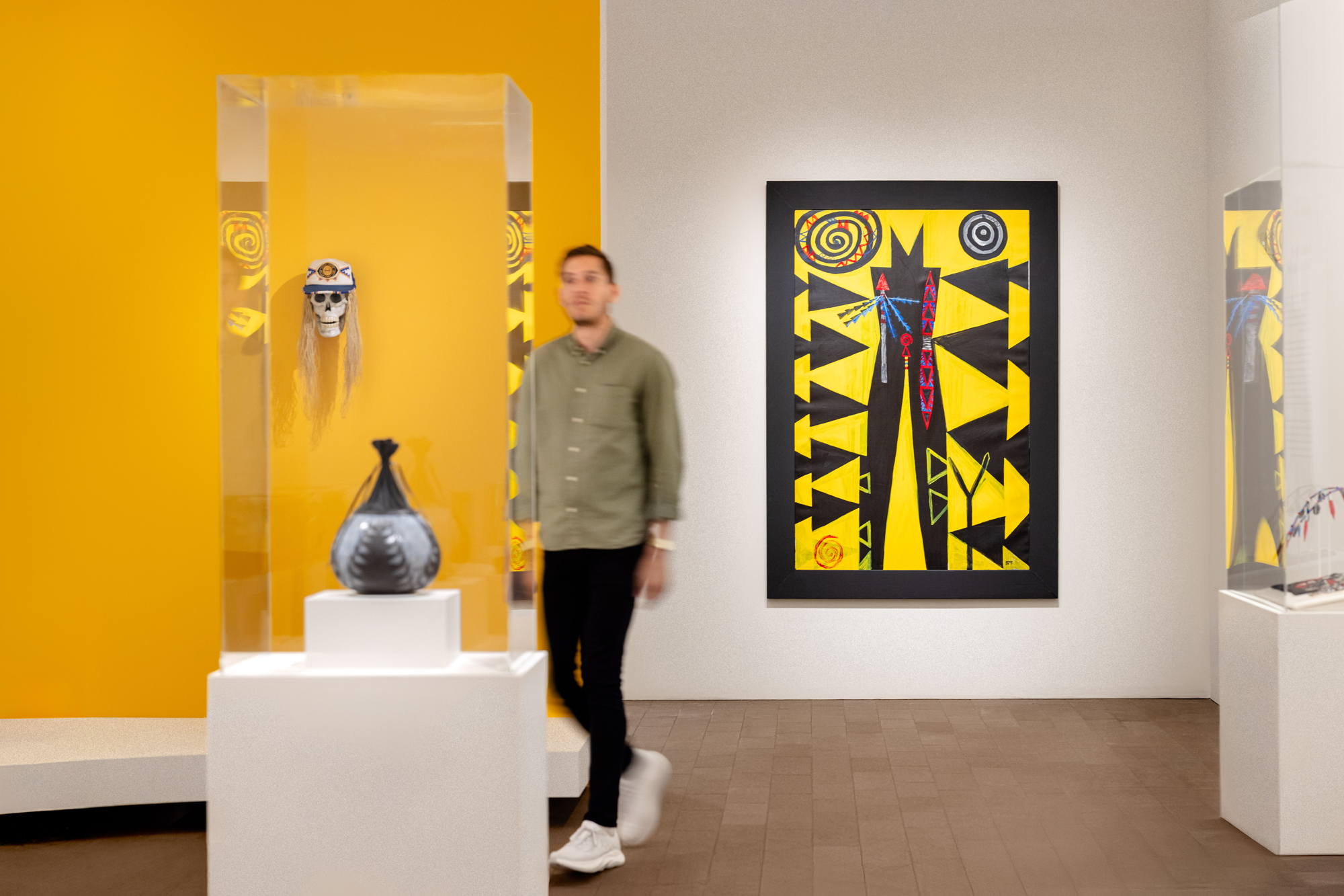 gallery shot with bright yellow wall, hanging art and sculptures in vitrines