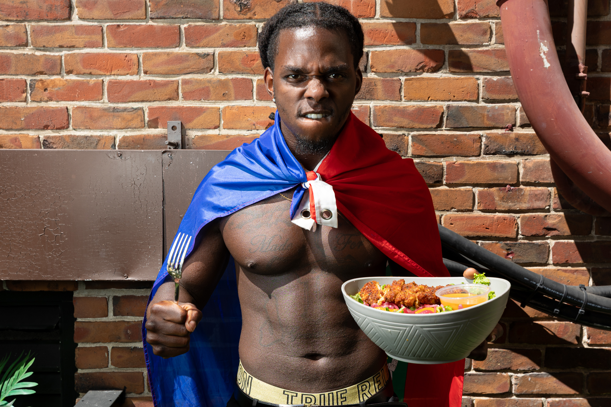 A Black man, shirtless besides a blue and red superhero cape, poses with a fierce expression while holding a bowl of salad.