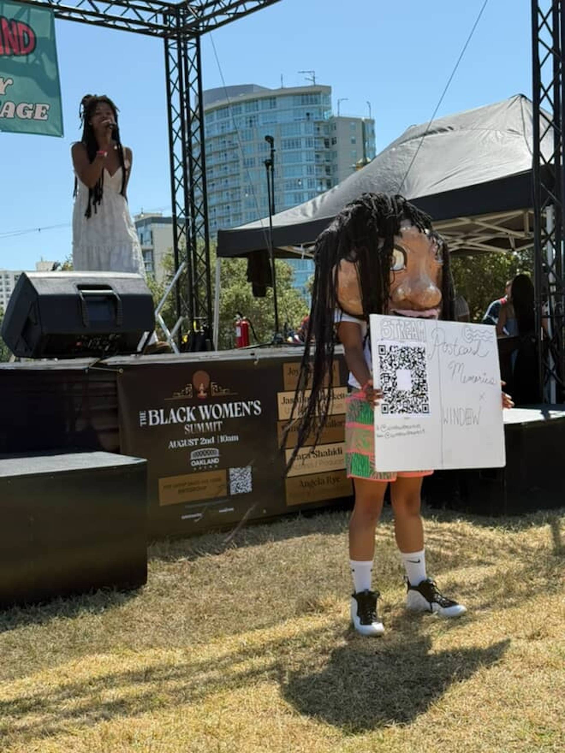 A person wears an oversized masked while standing in front of a stage where another person performs. 