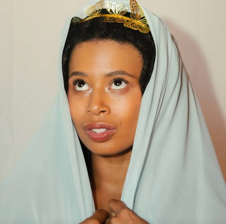 A Black woman looks up while wearing a white cloth and a golden crown on her head.