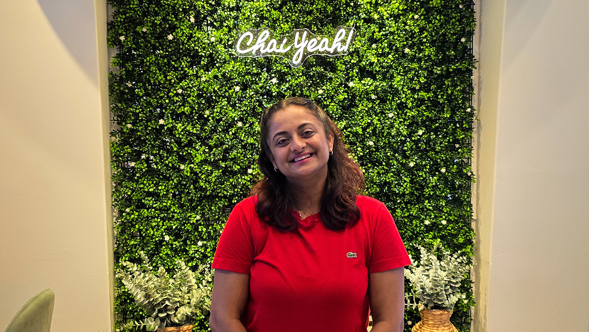 A woman poses in front of a green plant wall with a neon sign that reads, "Chai yeah."