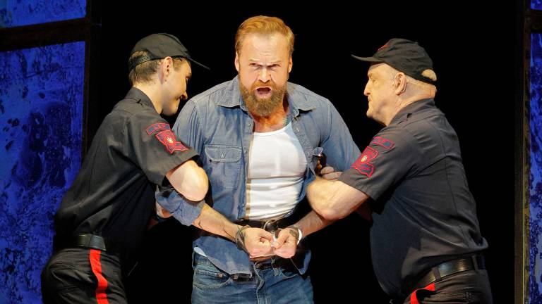 A man in denim shirt over a white t-shirt and in handcuffs sings while being led by two prison guards in uniform