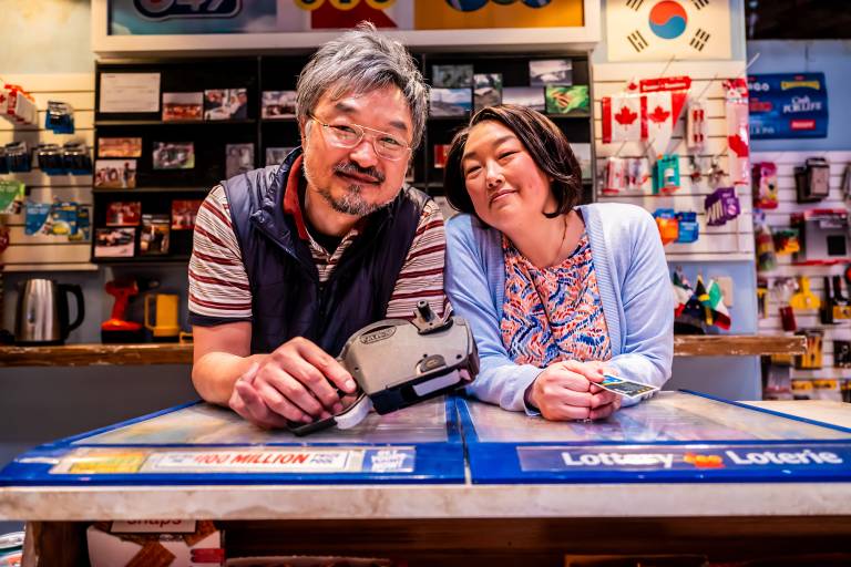 A Korean man and woman lean on a convenience store counter, the man holding a pricing gun, against a backdrop of candy and snacks