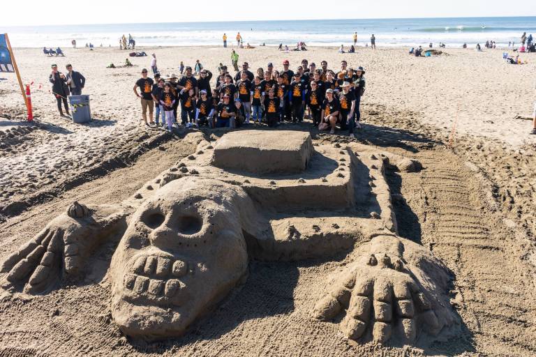 A giant sand sculpture of a temple with attached skull and hands sits on a bright beach with a large group of people standing behind it in matching t-shirts.