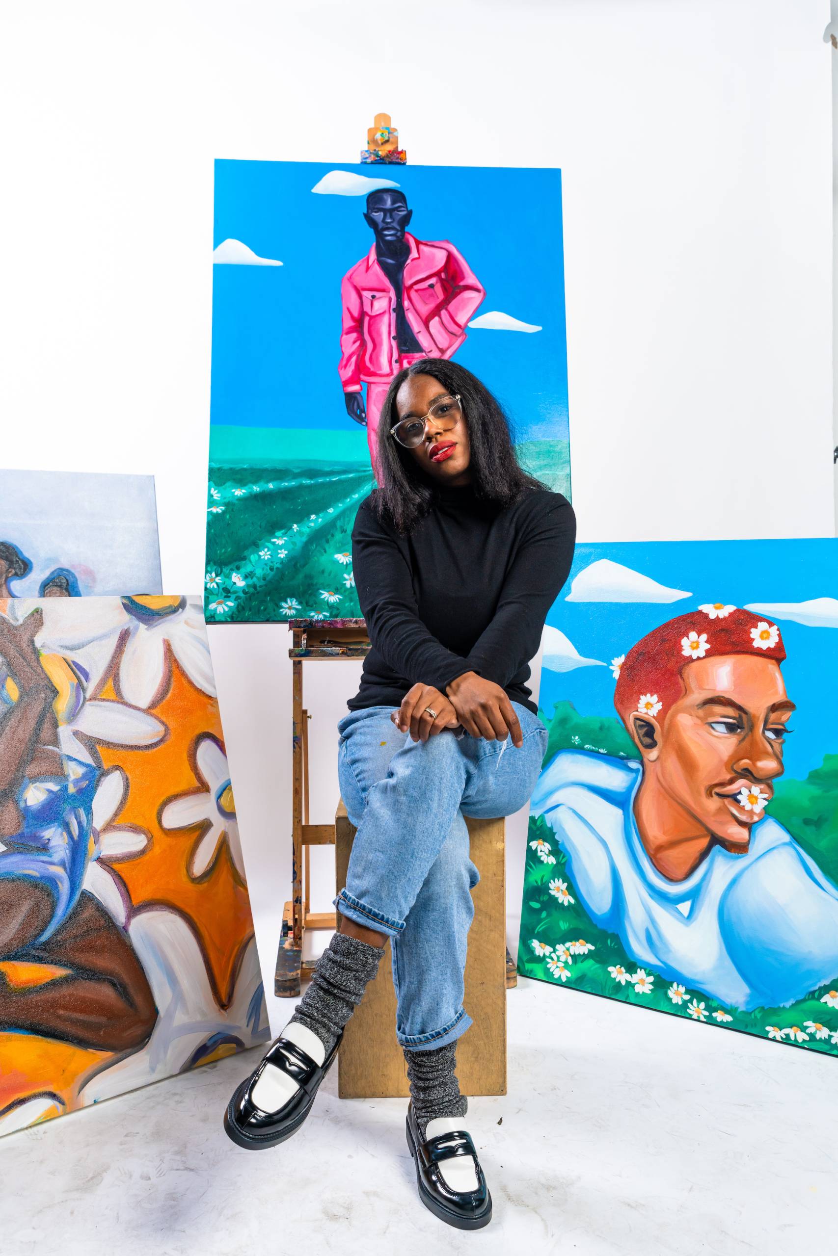 An African American woman wearing a black top, blue jeans and glasses, sitting in a studio with white walls surrounded by colorful paintings.