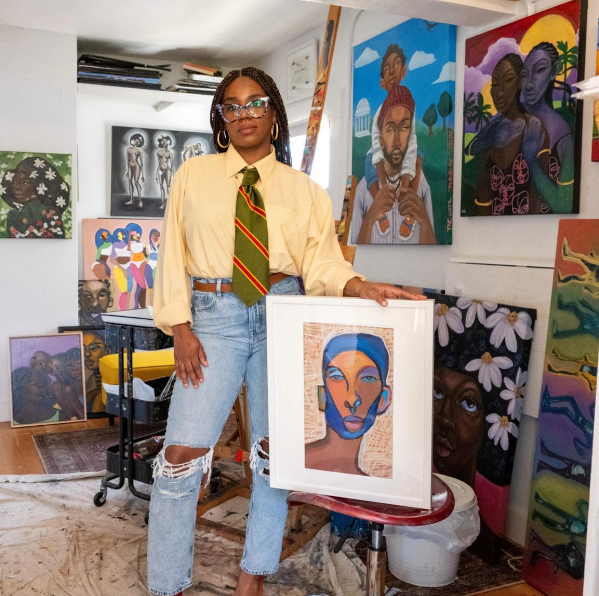 A woman in a yellow dress shirt, green and orange colored tie and blue jeans, stands in a room surrounded by her paintings. 