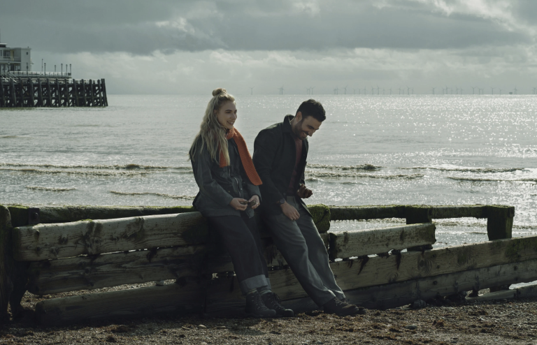 A woman and a man in warm clothes lean on a wooden platform on a beach.
