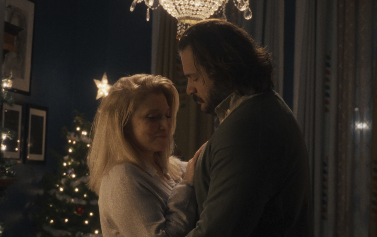 A tall white man with shaggy brown hair, wearing a green sweater, slow dances with a white blonde woman wearing a white sweater. She is looking down coyly.
