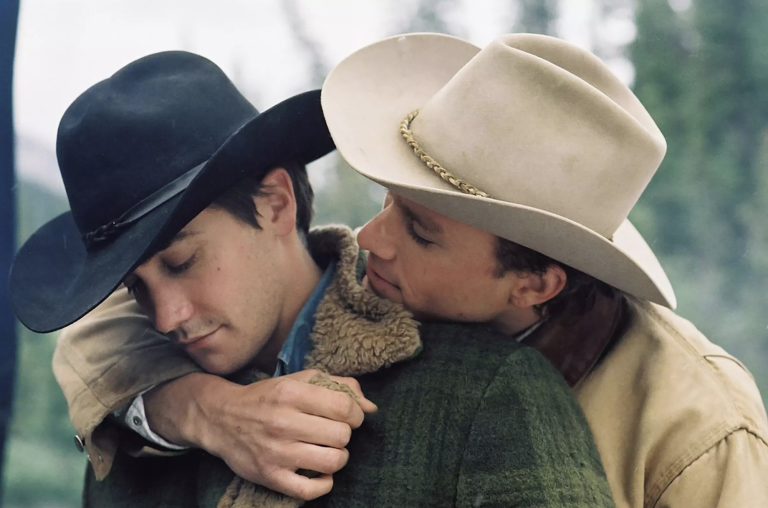 Two men wearing cowboys hats embrace tenderly in the outdoors.