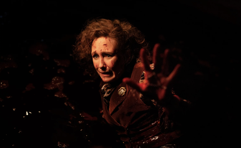 A disturbed looking woman with bloody flecks on her face stands in the dark with one arm raised.