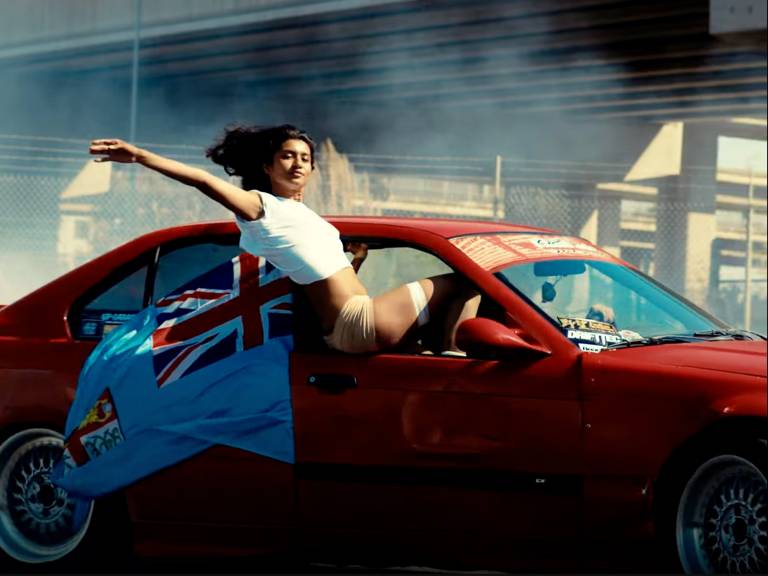 A woman in a white top hangs out the window of a red BMW car, leaning back with her right arm in the air as smoke emerges from the car's tires