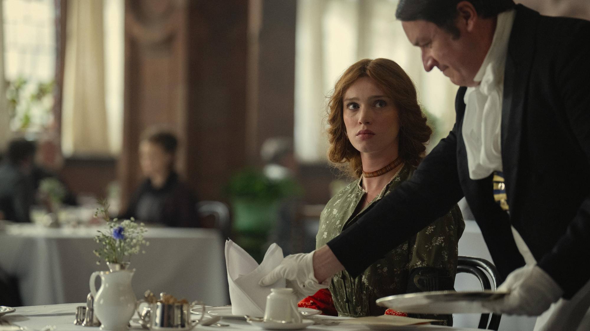 A young white woman in corseted period clothing is served afternoon tea in an upscale dining room.