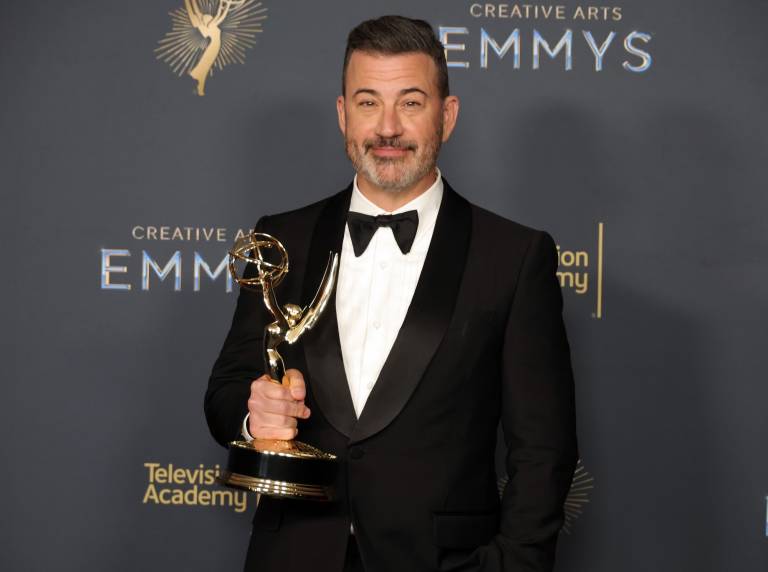 A middle aged white man with brown hair stands on a red carpet holding an Emmy Award. He is wearing a tuxedo.