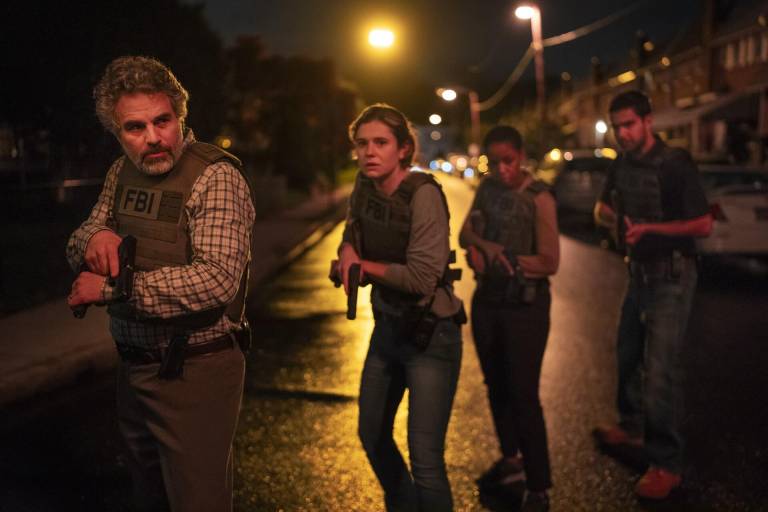 A white middle aged man wearing an FBI vest stands in the street at night, gun drawn, with three young people hovering behind him in similar stances.